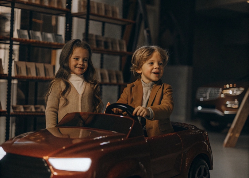 one girl and one boy about 5 years old standing next to a shiny red sedan in a modern ride on car showroom, smiling as they examine the vehicle. The background includes shelves with car paint samples and other cars slightly blurred. Bright and professional lighting fills the scene.